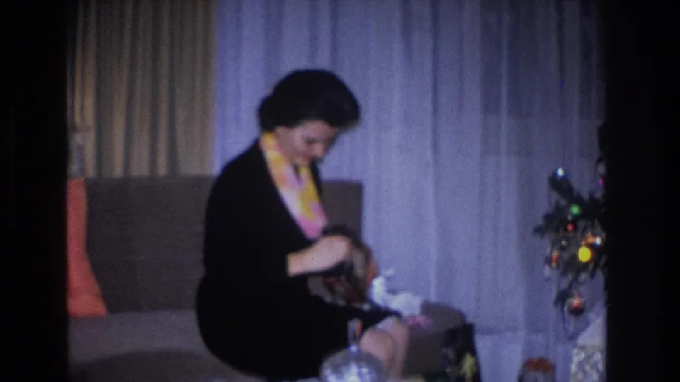 Woman in black dress and yellow scarf examining gift contents in her lap; Christmas tree with round multicolored ornaments at right; living room interior, circa 1966, Hoboken, NJ.