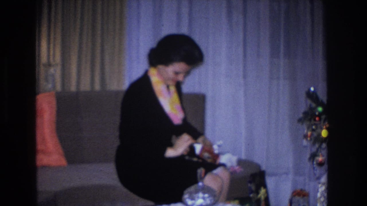 Woman in 1960s-style black dress and colorful scarf unwrapping a gift in a domestic living room beside a decorated Christmas tree, Hoboken NJ, 1966 home movie.