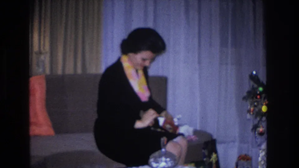 Woman in 1960s-style black dress and colorful scarf unwrapping a gift in a domestic living room beside a decorated Christmas tree, Hoboken NJ, 1966 home movie.