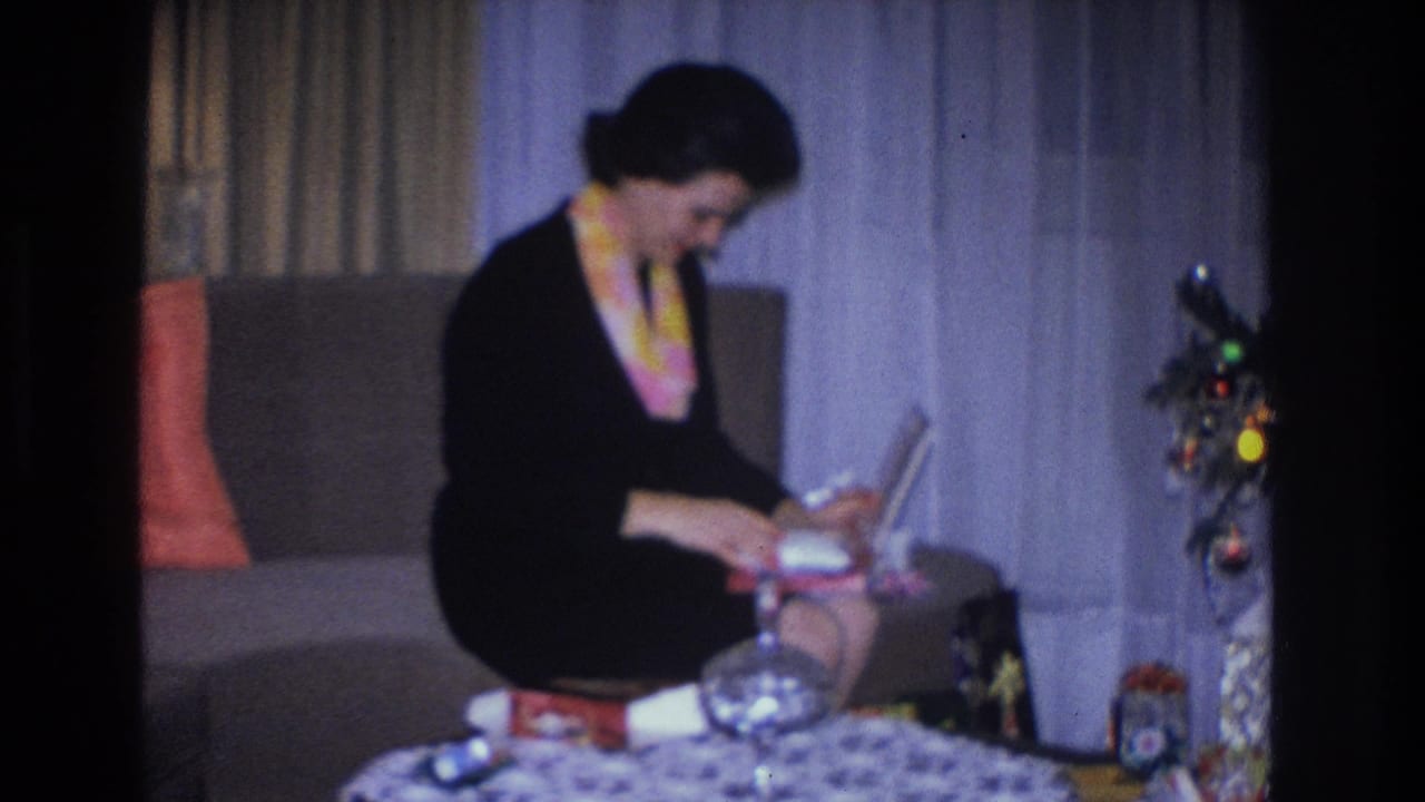 Woman holding a boxed gift while seated at a low table; Christmas tree with ball ornaments visible at right.