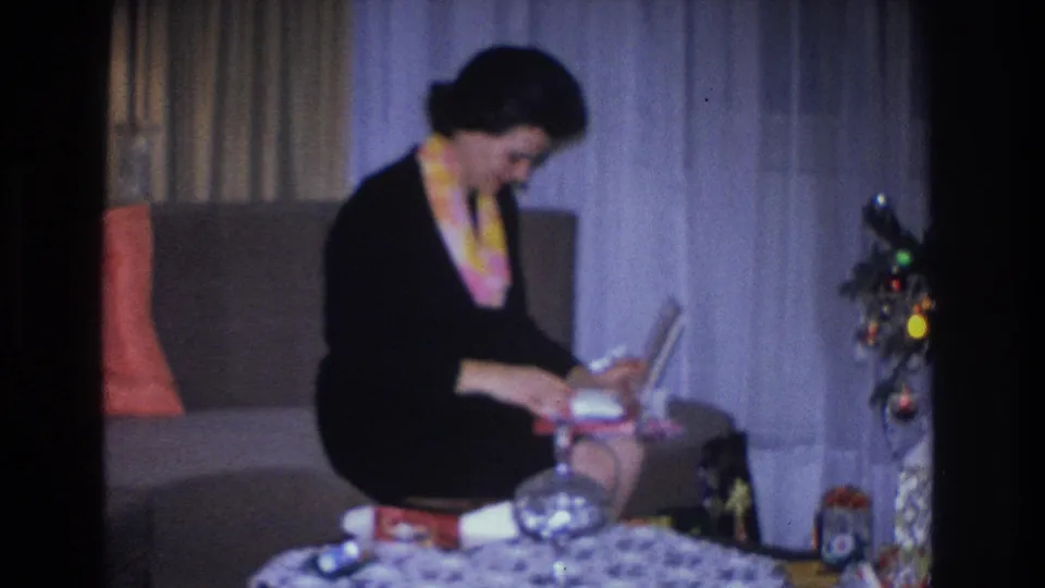 Woman in black dress examining a boxed Christmas gift in a mid-1960s living room, with decorated tree and patterned tablecloth, Hoboken NJ home movie.