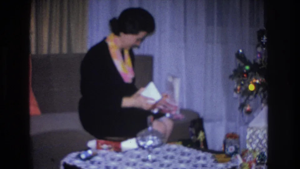 Woman in 1960s dress actively unwrapping a present beside a small Christmas tree, with glass bowl and stacked gifts on patterned tablecloth, home movie.