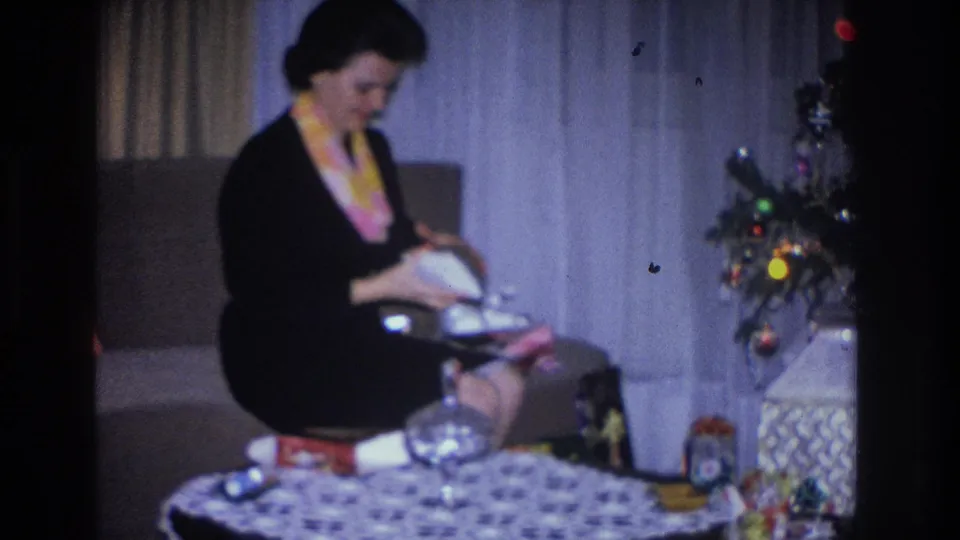 Woman inspecting a round gift item in a mid-1960s living room decorated for Christmas, with tree, wrapped packages, and patterned tablecloth visible, Hoboken NJ home movie.