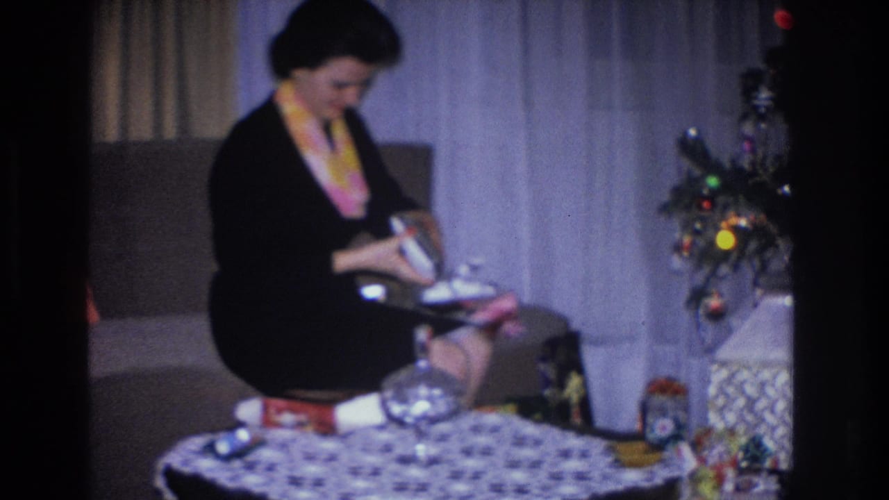 Woman in black dress examines a gift box while seated on a couch beside a decorated Christmas tree