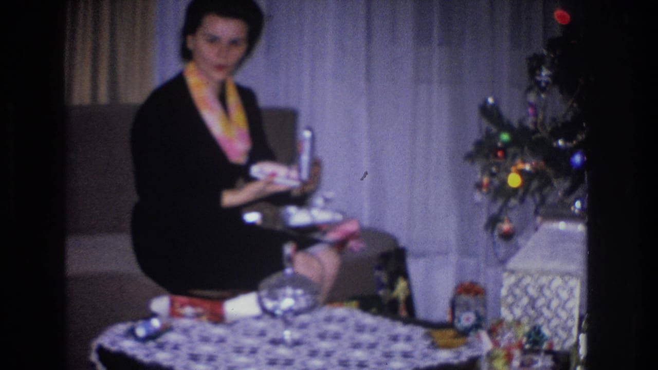 A woman in a black dress and yellow scarf holds a cylindrical gift upright while seated beside a decorated Christmas tree in a 1966 Hoboken, New Jersey home movie
