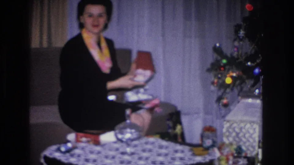 A woman in a black dress and yellow scarf inspects an open gift box beside a Christmas tree with colored ornaments in a 1966 Hoboken, New Jersey home movie