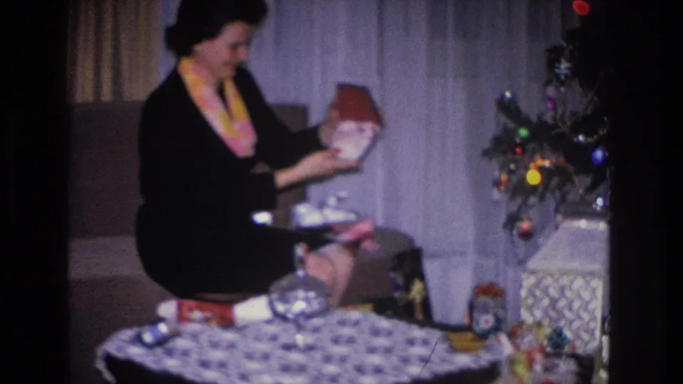 A woman in a black dress and yellow scarf holds a red-wrapped gift beside a decorated Christmas tree in a 1966 Hoboken, New Jersey home movie