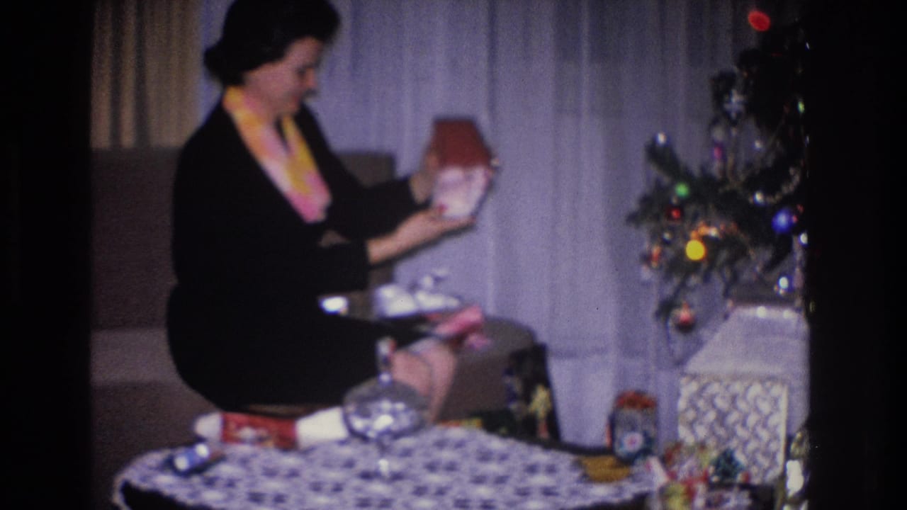 A woman in a black dress with a colorful scarf unwraps a Christmas gift in what appears to be a 1966 Hoboken, New Jersey home, with a decorated tree visible behind her.
