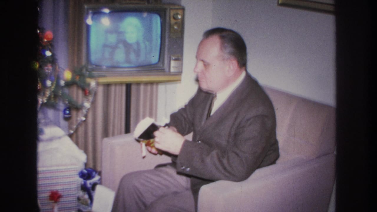 Man in dark suit opens a gift in an armchair; a vintage television with a glowing screen and decorated Christmas tree are visible behind him.