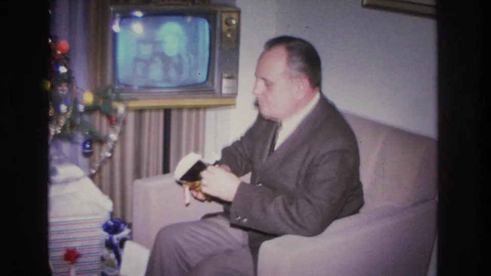 A man in a dark suit and tie unwraps a Christmas gift in an armchair in what appears to be a 1966 Hoboken home, with a vintage television set and decorated tree in the background.