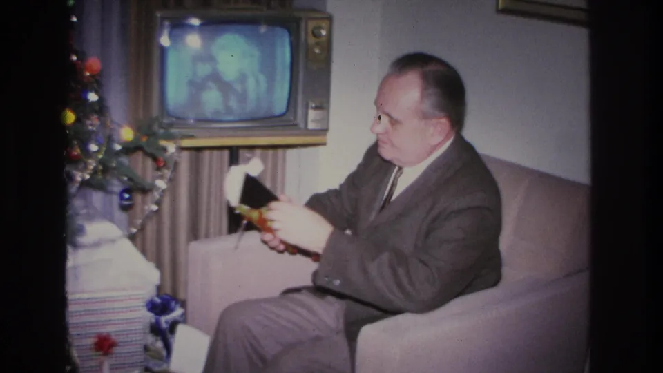 The same man in a dark suit continues opening a Christmas gift in an armchair, with colored tree ornaments and a glowing vintage television prominent to his left, circa 1966.