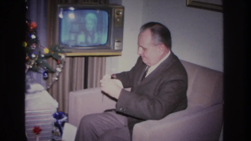 In the final frame of this 1966 home movie, the man in a dark suit continues unwrapping a gift, with the Christmas tree and glowing vintage television still visible behind him.