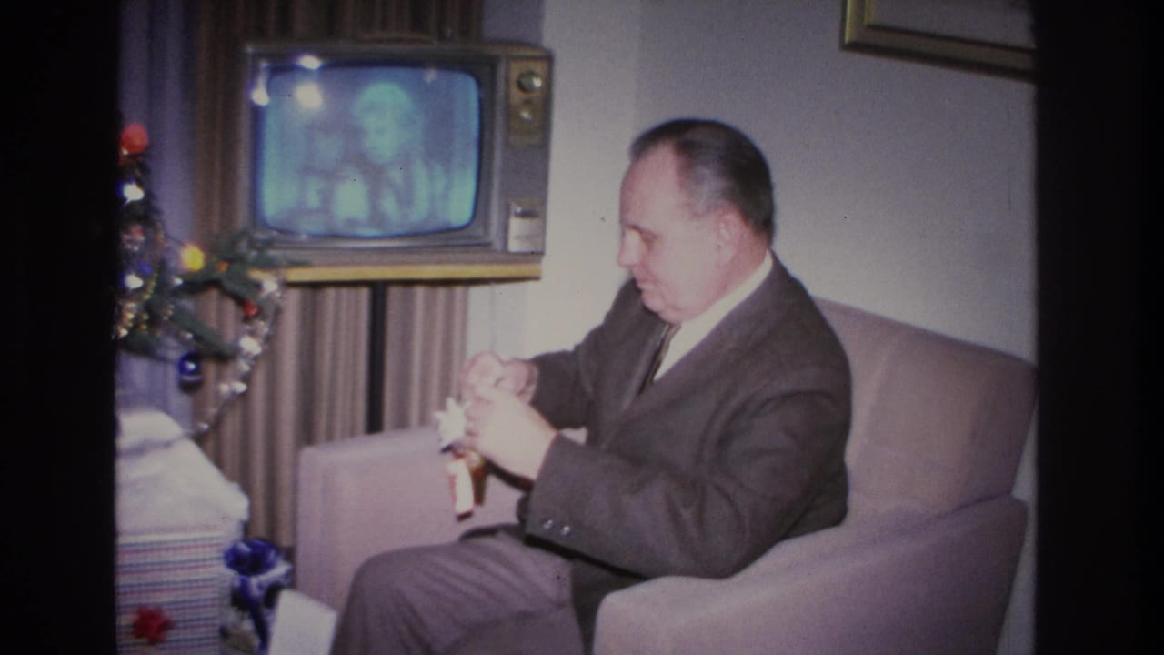 Middle-aged man in dark suit begins unwrapping a Christmas gift in an armchair in a 1966 Hoboken, New Jersey home; wood-cabinet console TV and decorated tree visible behind him.