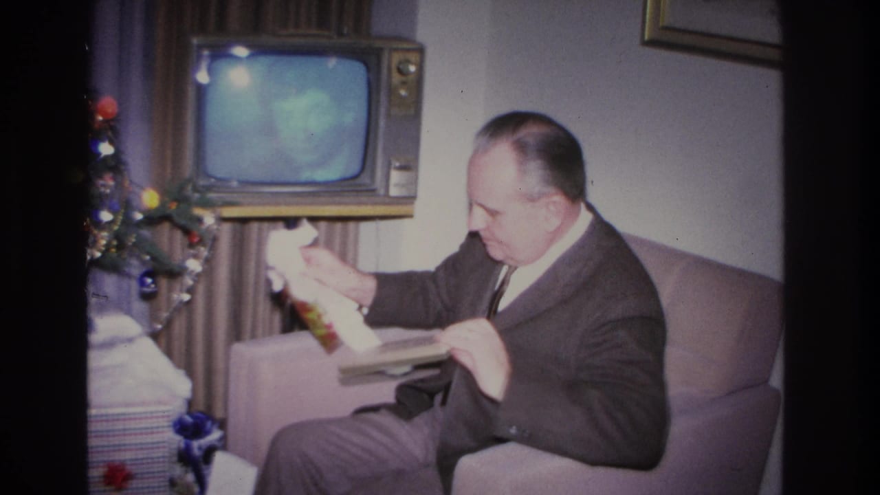 Man in dark suit holds up a flat book or album retrieved from gift wrapping; wood-cabinet console TV and Christmas tree visible in background.