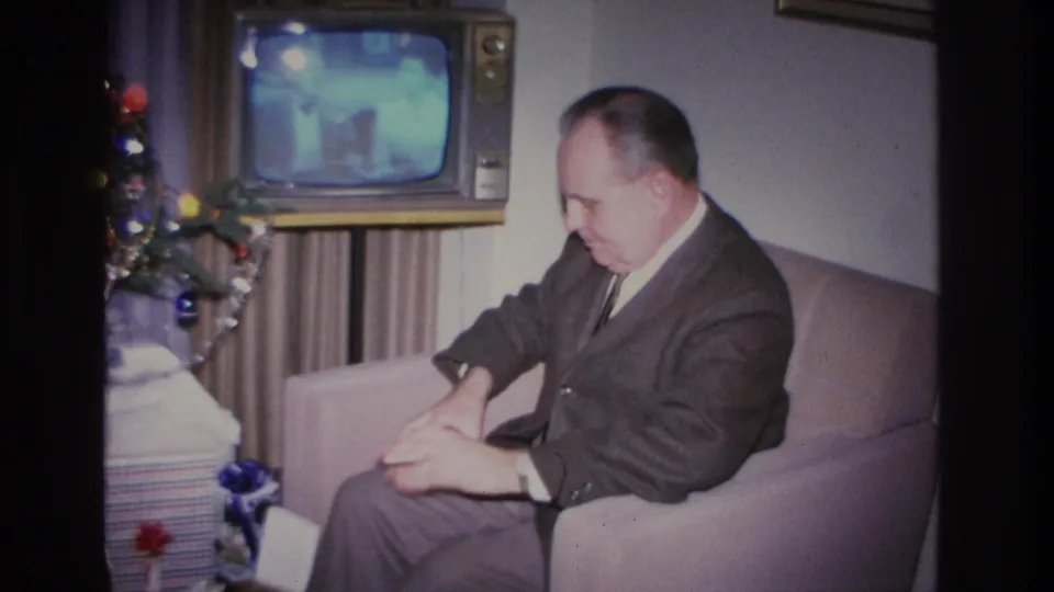 Man in dark suit looking down at a gift in his lap in a 1966 living room interior; vintage console TV still broadcasting and Christmas tree with colorful ornaments at frame left.