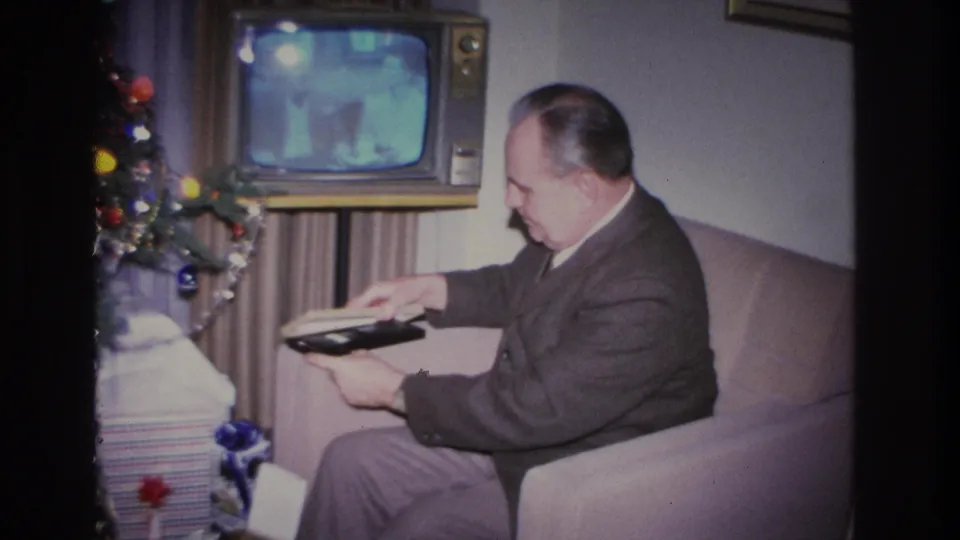 Man in dark suit holds and examines a dark-covered book or album in a 1966 Hoboken, New Jersey living room; vintage console TV in background, Christmas tree with colorful ball ornaments at left.