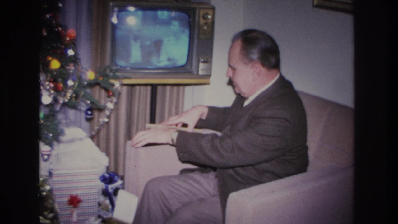 Middle-aged man in dark suit seated on a light sofa, with a vintage console television on and a decorated Christmas tree visible to the left.