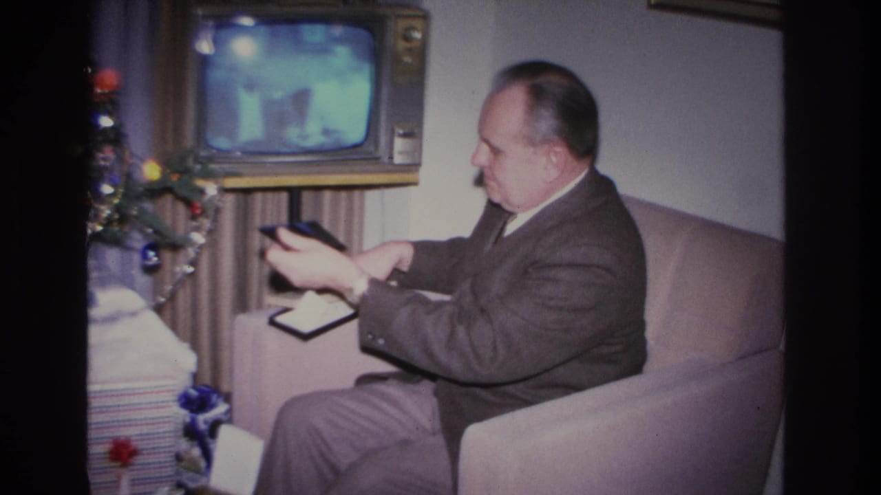 Man in a 1960s suit holds a small dark rectangular object, possibly a remote control, while seated on a sofa; vintage console TV on and decorated Christmas tree at left, Hoboken New Jersey 1966.
