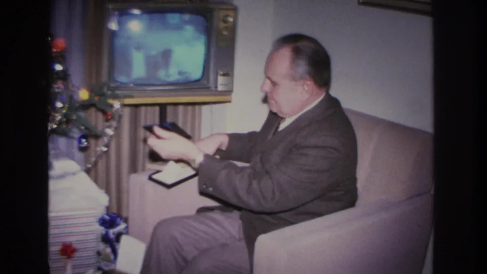 Man in a 1960s suit holds a small dark rectangular object, possibly a remote control, while seated on a sofa; vintage console TV on and decorated Christmas tree at left, Hoboken New Jersey 1966.