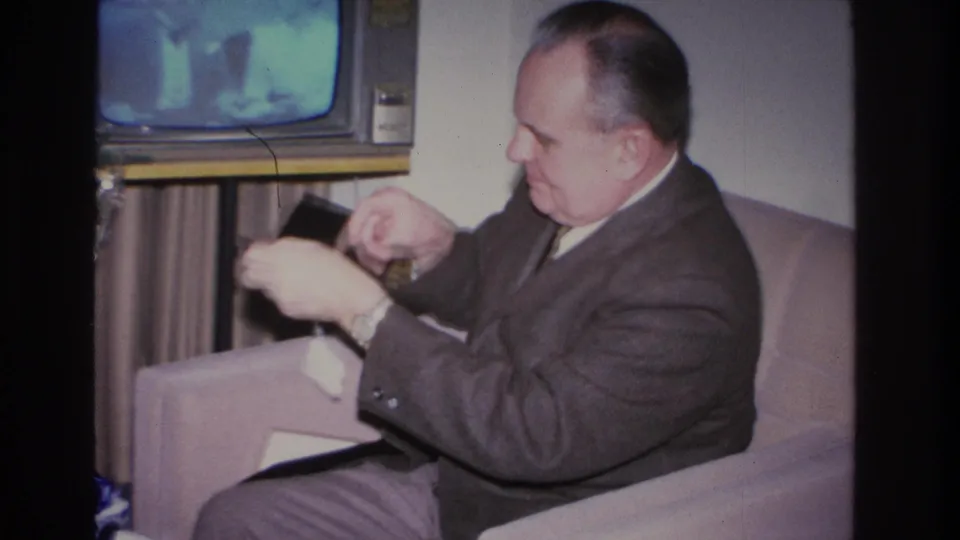 Man in a 1960s suit leans forward on sofa, inspecting a small dark object at close range; console television and partial Christmas tree in background, Hoboken New Jersey 1966.