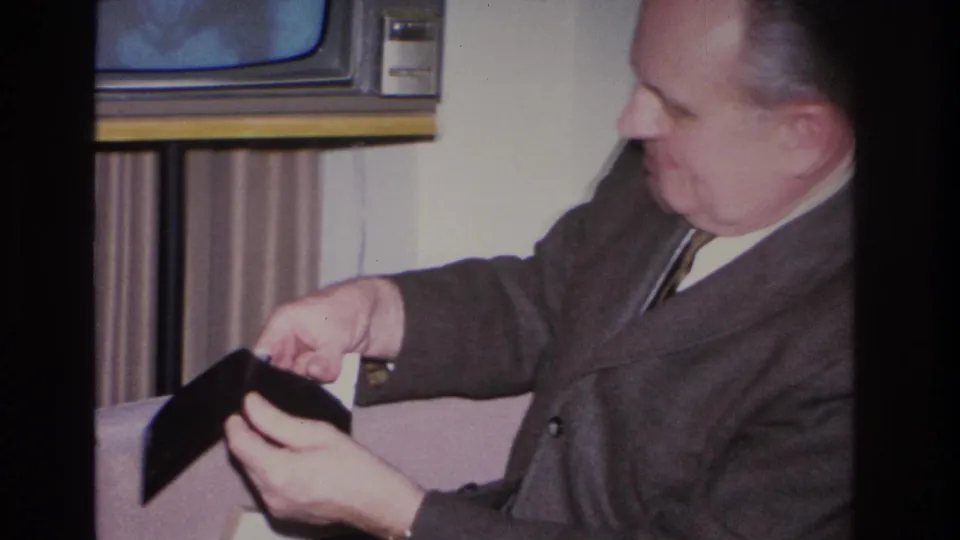 Close shot of man in a 1960s suit holding a small dark rectangular object, possibly a remote control or small case; vintage television partially visible behind, Hoboken New Jersey 1966.