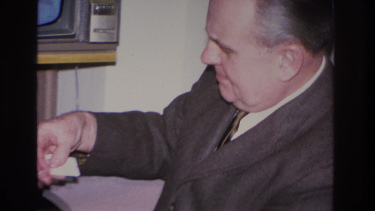 Middle-aged man in dark suit leaning forward with arm extended, vintage CRT television visible in upper-left background corner.