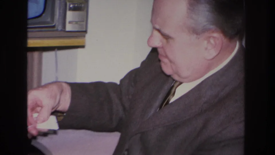 Middle-aged man in dark suit and narrow tie leans forward with arm extended in a domestic interior; period CRT television visible in background; Hoboken, New Jersey, 1966 per metadata.