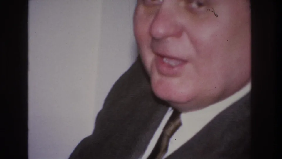 Close frontal view of middle-aged man in dark suit showing an animated, open-mouthed expression; a film scratch runs across the frame; home movie footage circa 1966.