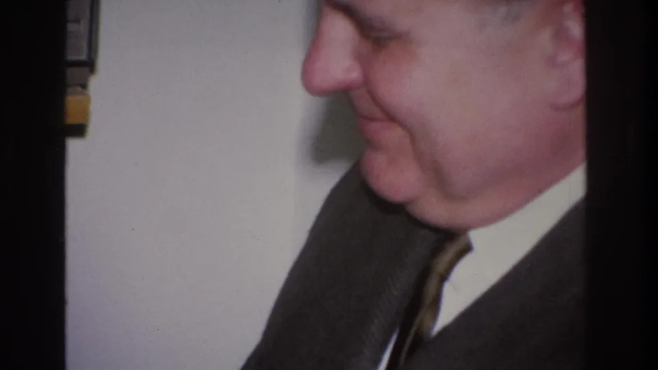 Close-up side profile of a middle-aged man in a dark charcoal suit and striped tie, 1966 8mm home movie, Hoboken, New Jersey.
