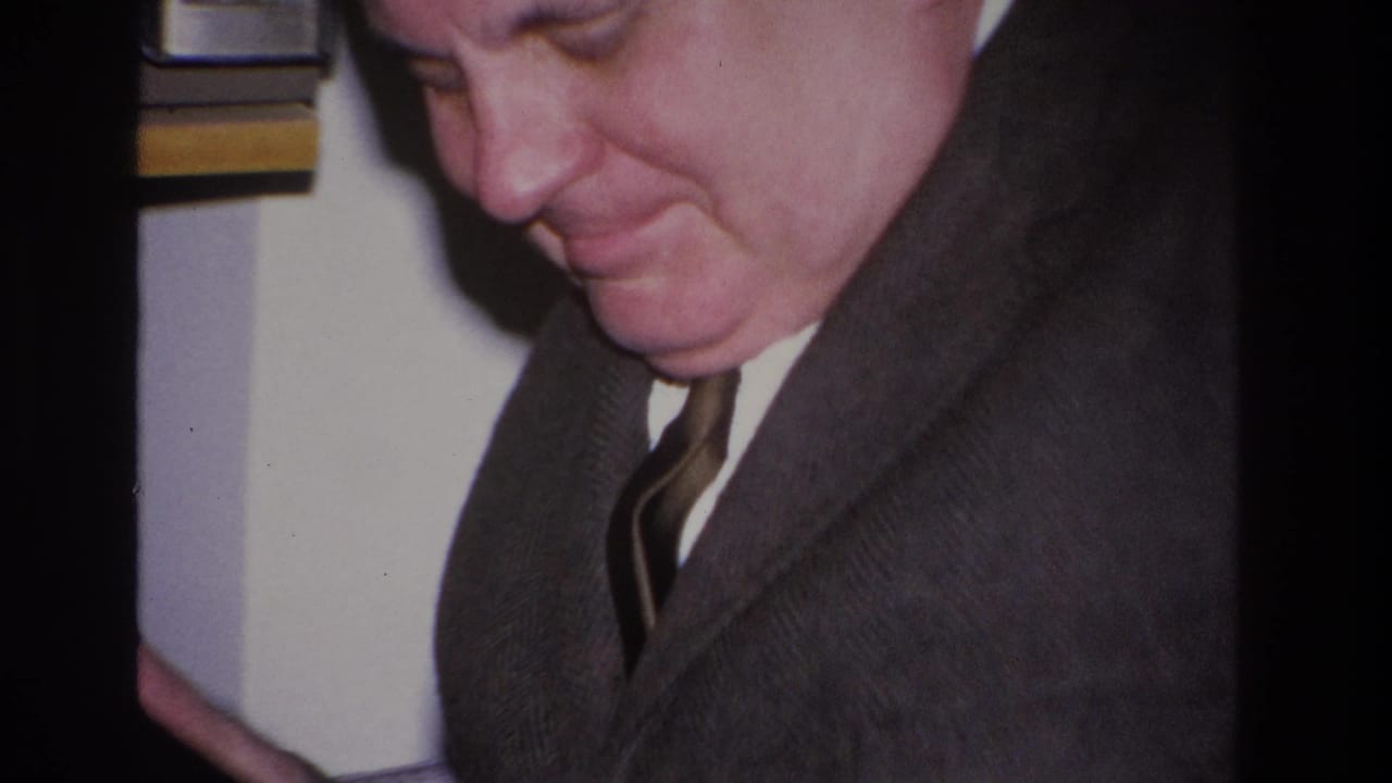 Three-quarter profile of a middle-aged man in a dark suit and striped tie, smiling gently while looking downward.