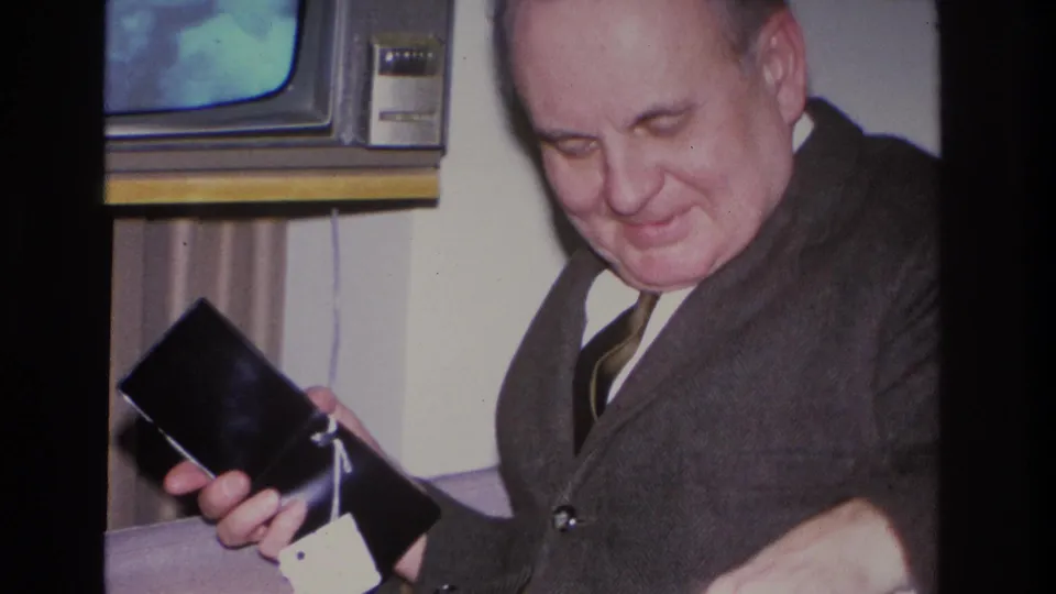 Middle-aged man in dark suit holding a small black case with a white tag, smiling in a domestic living room with a 1960s console television behind him; 1966 8mm home movie, Hoboken, NJ.