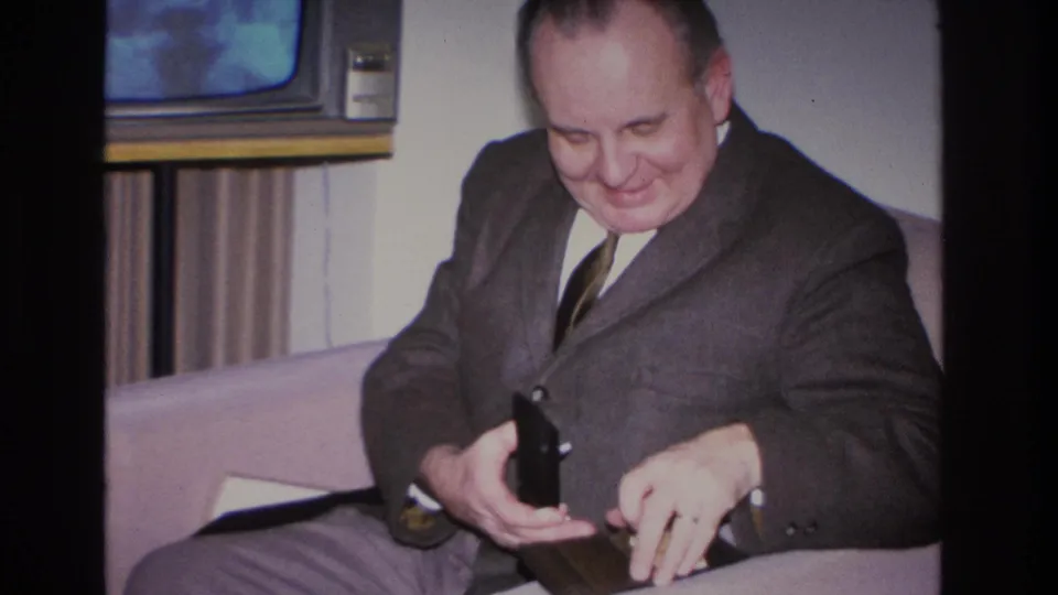 Middle-aged man in dark suit operating or examining a small black case while seated on a sofa, 1960s console television with glowing screen in background; 1966 8mm home movie, Hoboken, NJ.