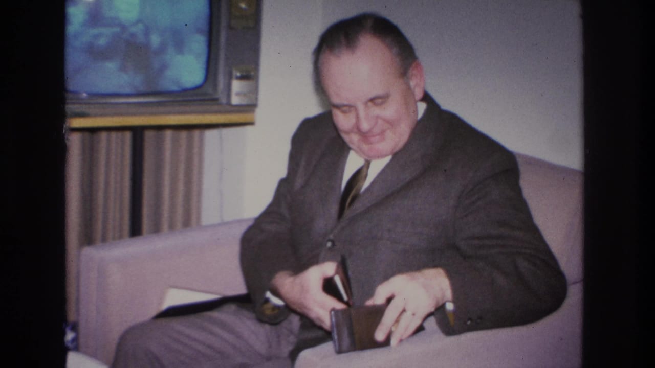 Middle-aged man in dark suit smiling as he opens a dark wallet on a light sofa; boxy vintage television with blue-glowing screen visible behind him.