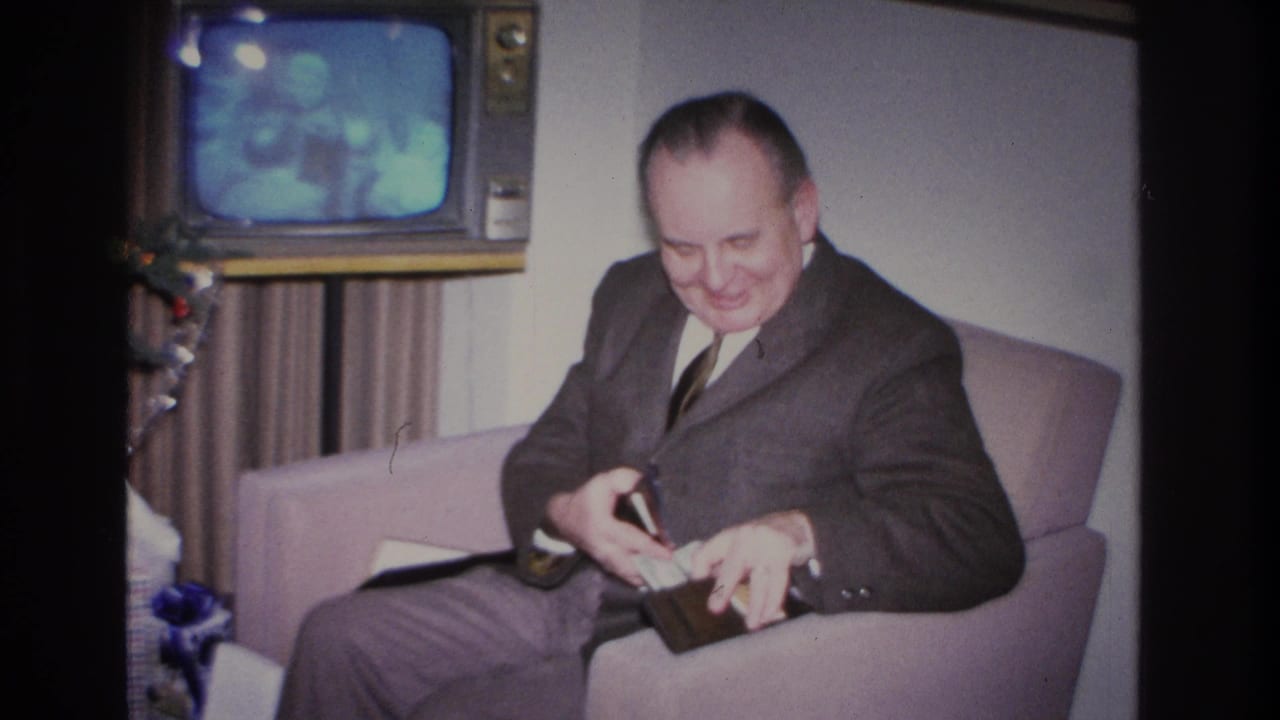 Middle-aged man in 1960s dark suit handling open wallet on sofa; boxy mid-century television visible upper right, decorated Christmas tree with blue ornaments at right edge.