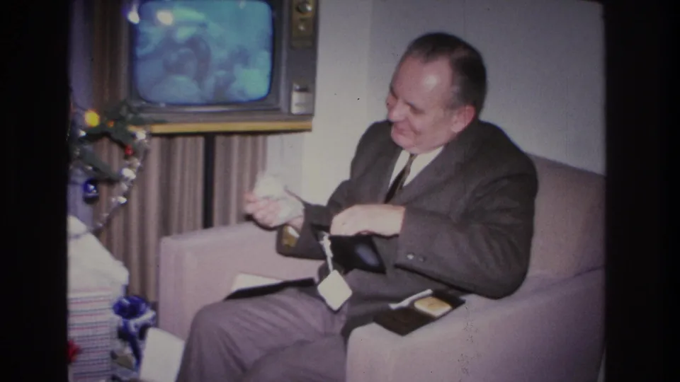 Middle-aged man in 1960s suit smiling broadly while opening wallet on lavender sofa; Christmas tree with blue ornaments and tinsel at left, boxy vintage television behind.