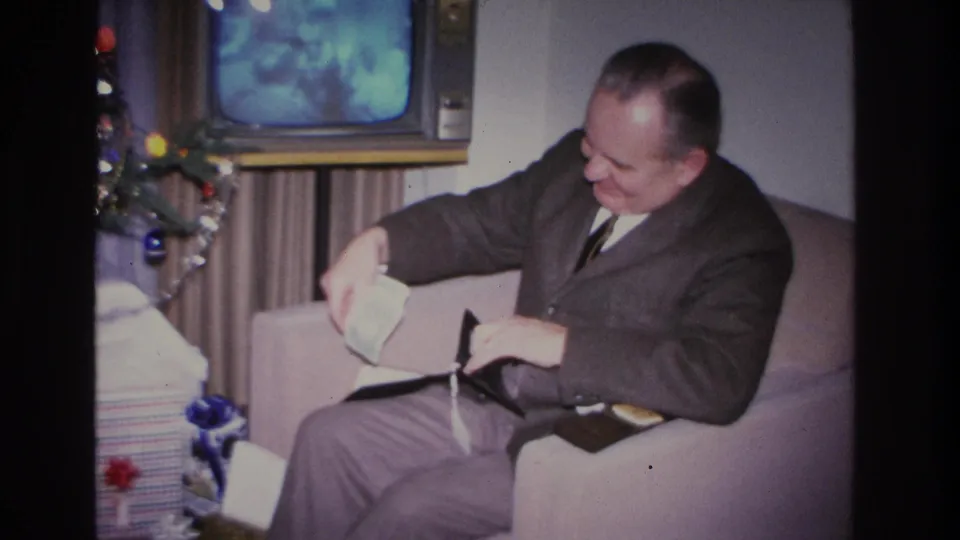 Middle-aged man in 1960s suit holding cash extracted from wallet while seated on lavender sofa; Christmas tree with blue ornaments and garland at right, vintage television behind.