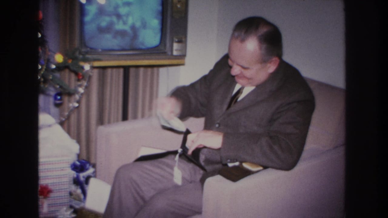 Middle-aged man in dark suit seated in armchair examining a wallet; decorated Christmas tree and vintage TV visible behind him.