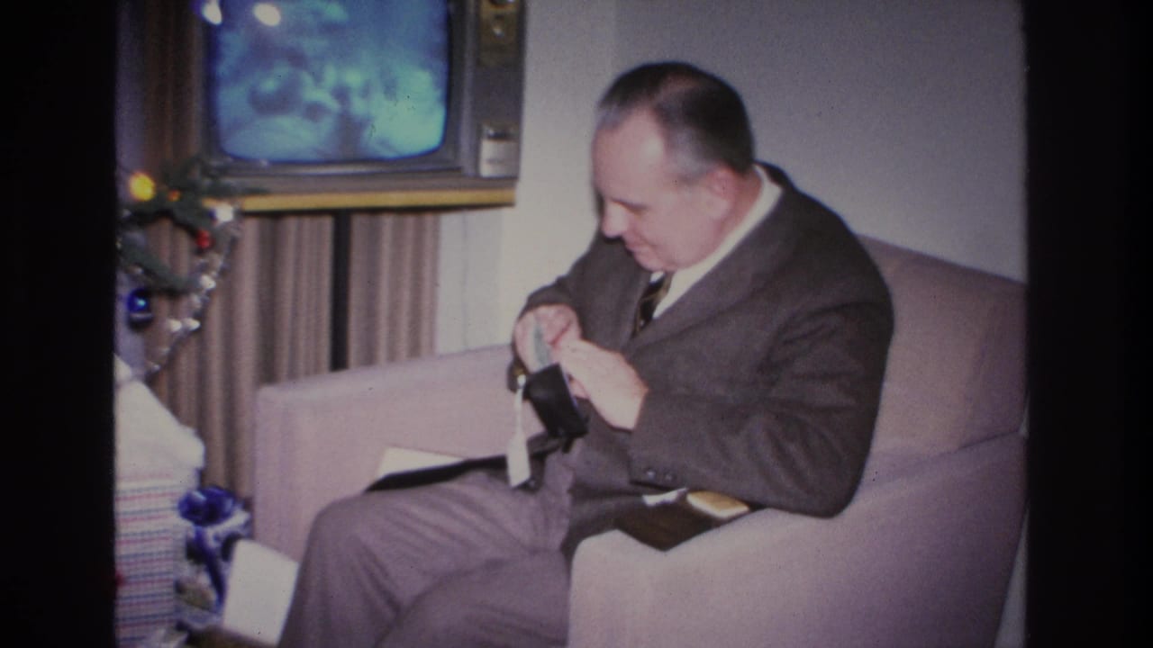 Man in mid-1960s suit holds wallet gift in domestic living room during Christmas; blue-screen TV and tree ornaments visible behind him.