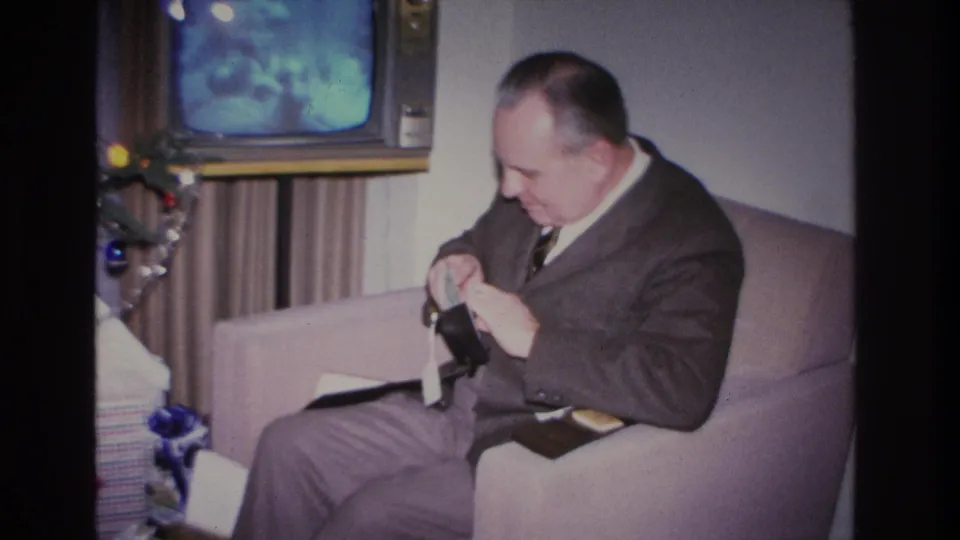 Man in mid-1960s suit holds wallet gift in domestic living room during Christmas; blue-screen TV and tree ornaments visible behind him.