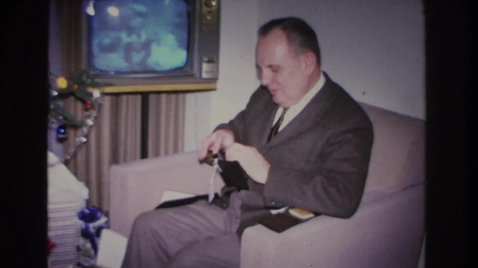 Man inserts a bill into a wallet gift in a 1966 domestic interior; vintage cathode-ray TV and decorated Christmas tree partially visible at left.