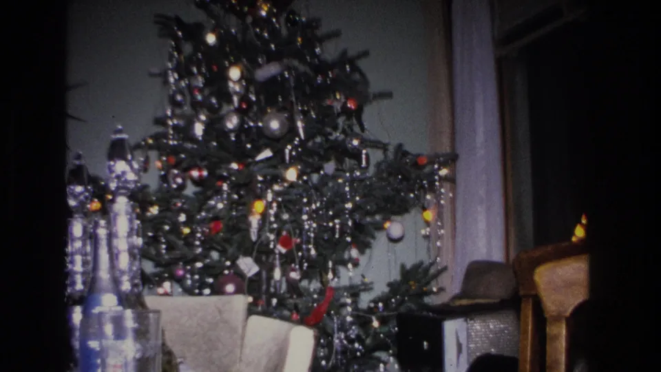 Decorated 1960s Christmas tree with colored ornaments, tinsel icicles, and string lights in a domestic living room; bottles and gifts at base; lamp visible at right.