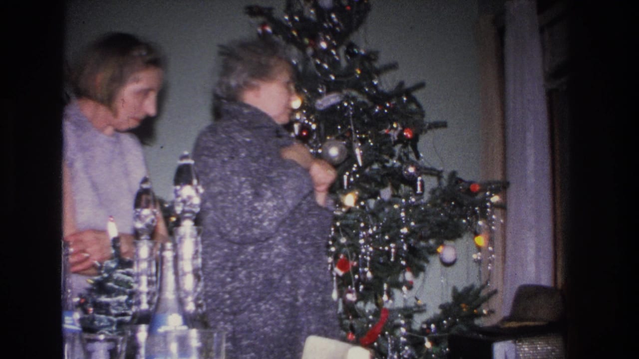 Two women seen in profile near a Christmas tree, with glass decanters and figurines on a table in the foreground.