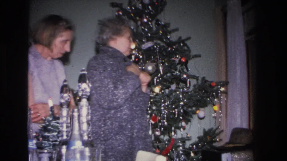 Two women in profile beside a decorated Christmas tree; the older woman wears a dark textured knit coat, in what appears to be a mid-1960s domestic interior.
