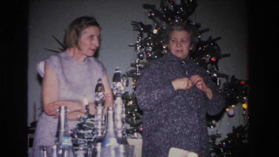 Younger woman in a sleeveless lavender dress and older woman in a dark knit coat face the camera in a mid-century living room decorated for Christmas.