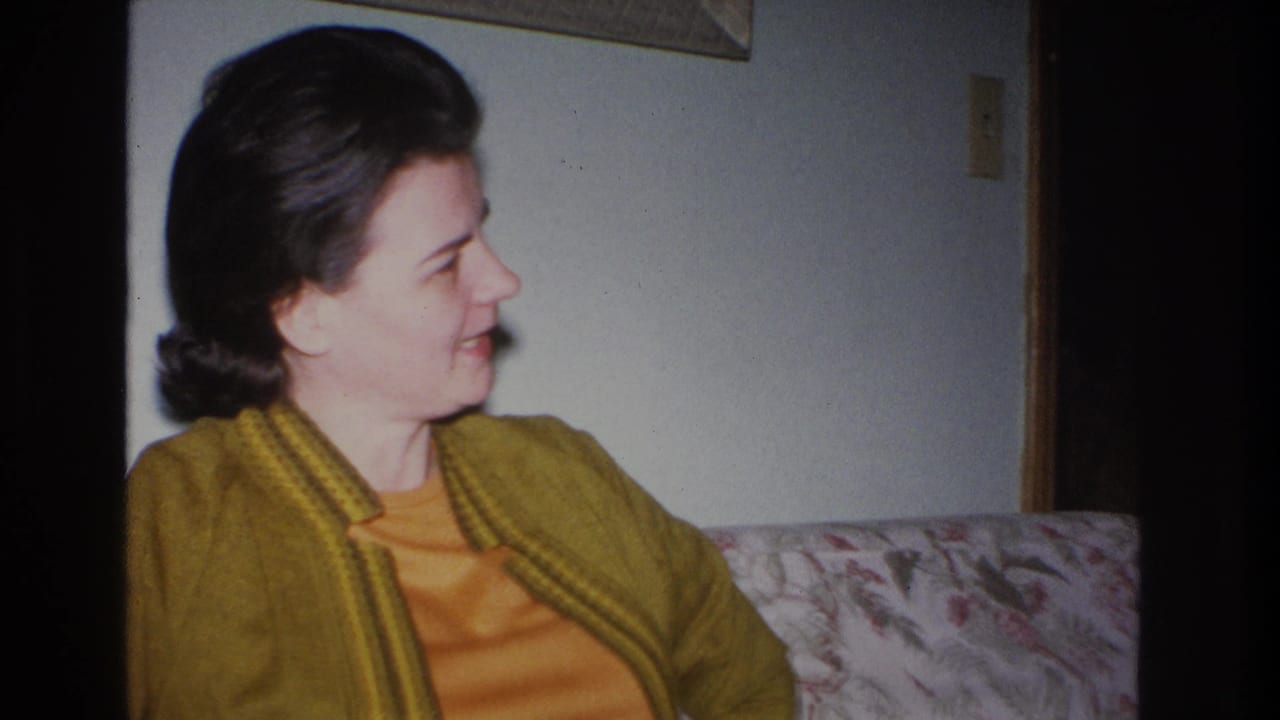 1960s woman in mustard cardigan over orange knit top seated on a floral-upholstered couch, smiling off-camera; 8mm home movie, likely Hoboken, New Jersey, 1966.