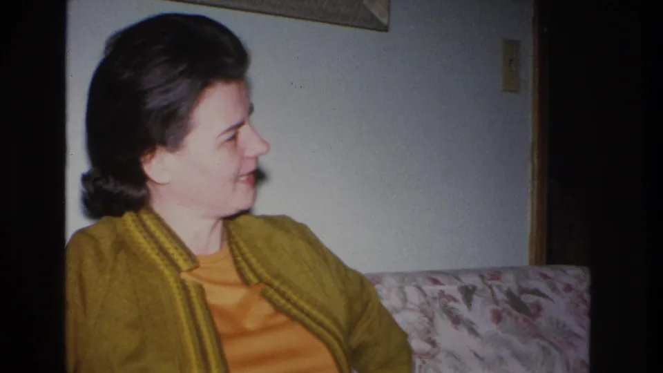 1960s woman in mustard cardigan over orange knit top seated on a floral-upholstered couch, smiling off-camera; 8mm home movie, likely Hoboken, New Jersey, 1966.