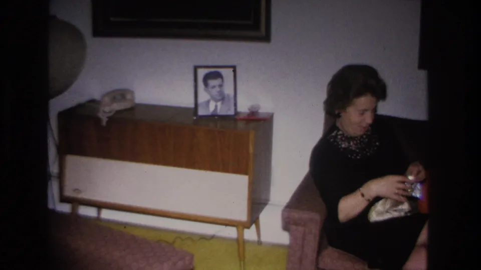 Woman in dark dress handling a small gift, seated beside a mid-century teak-and-white console bearing a framed male portrait and telephone; domestic interior consistent with Hoboken, New Jersey, c. 1966.