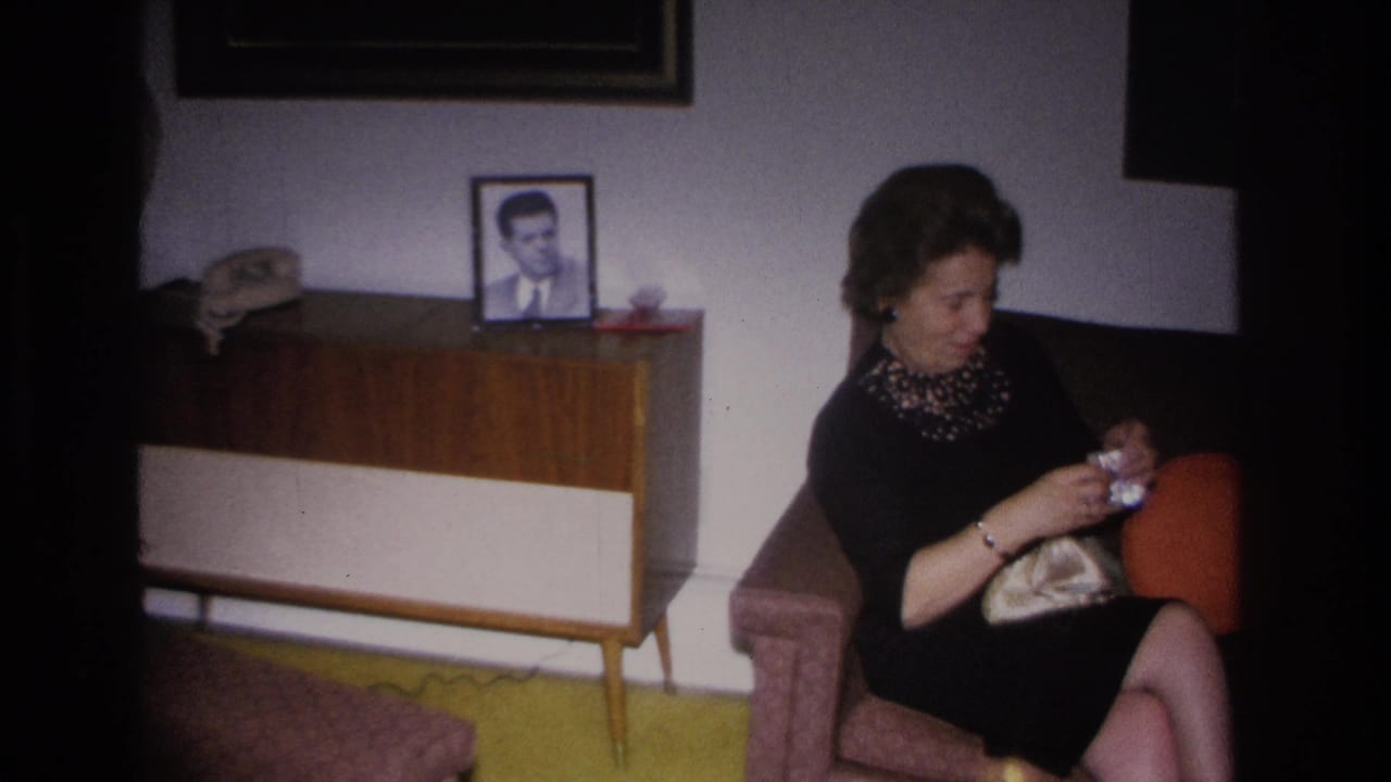 Mid-1960s woman in beaded black dress opens a clutch purse on an armchair; wood sideboard with a man's framed portrait and mustard-yellow carpet in background, Hoboken 1966.