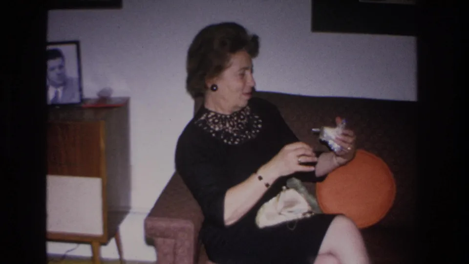 Mid-1960s woman in beaded black cocktail dress retrieves item from open clutch; orange round pillow and mid-century sideboard in background, Hoboken living room 1966.