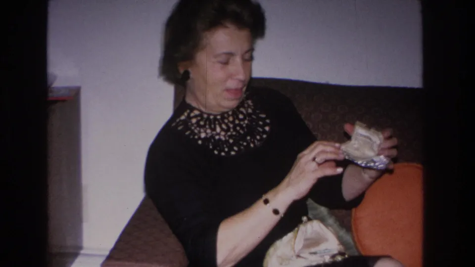 Mid-1960s woman in embellished black dress examines something retrieved from her clutch; orange round pillow and framed portrait remain visible in the Hoboken living room background.